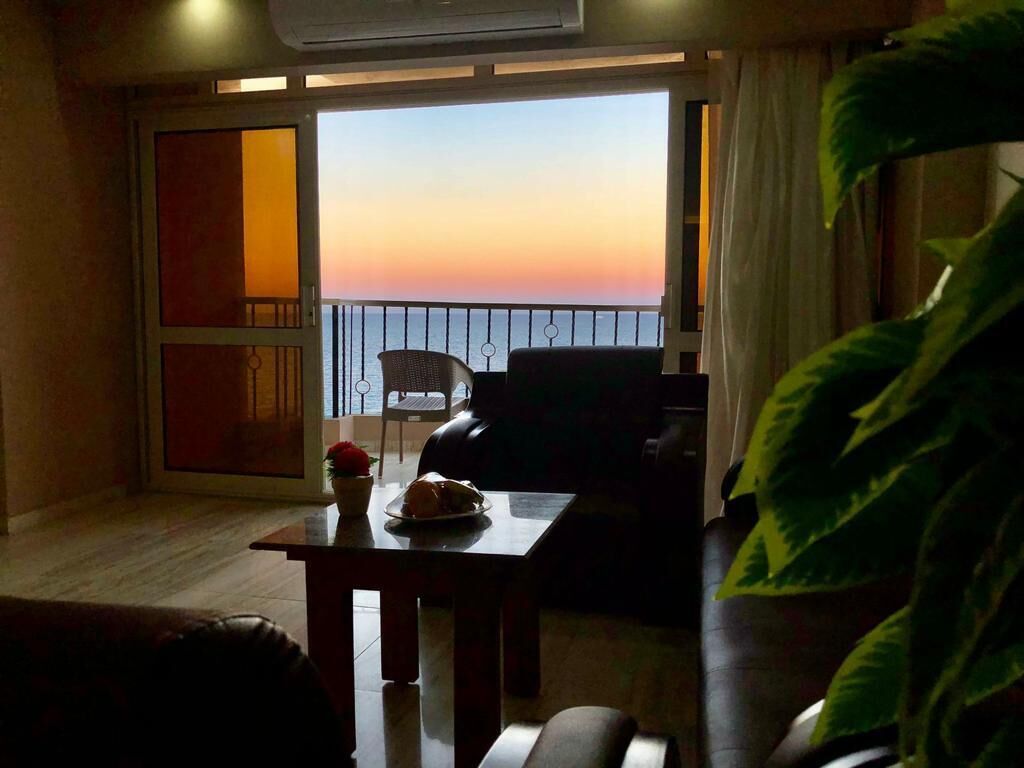 A room with a view of the sea at sunset, a dining table with a plate of food, a sofa, and a large green plant.