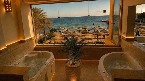 Une salle de bain avec deux jacuzzis, une plante en pot et une vue sur la plage, avec des parasols.