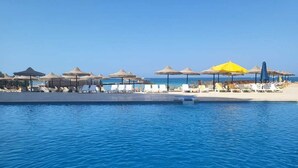 Un espace aménagé au bord de la piscine, avec des chaises longues et des parasols, un ciel bleu azur et une mer calme en arrière-plan.