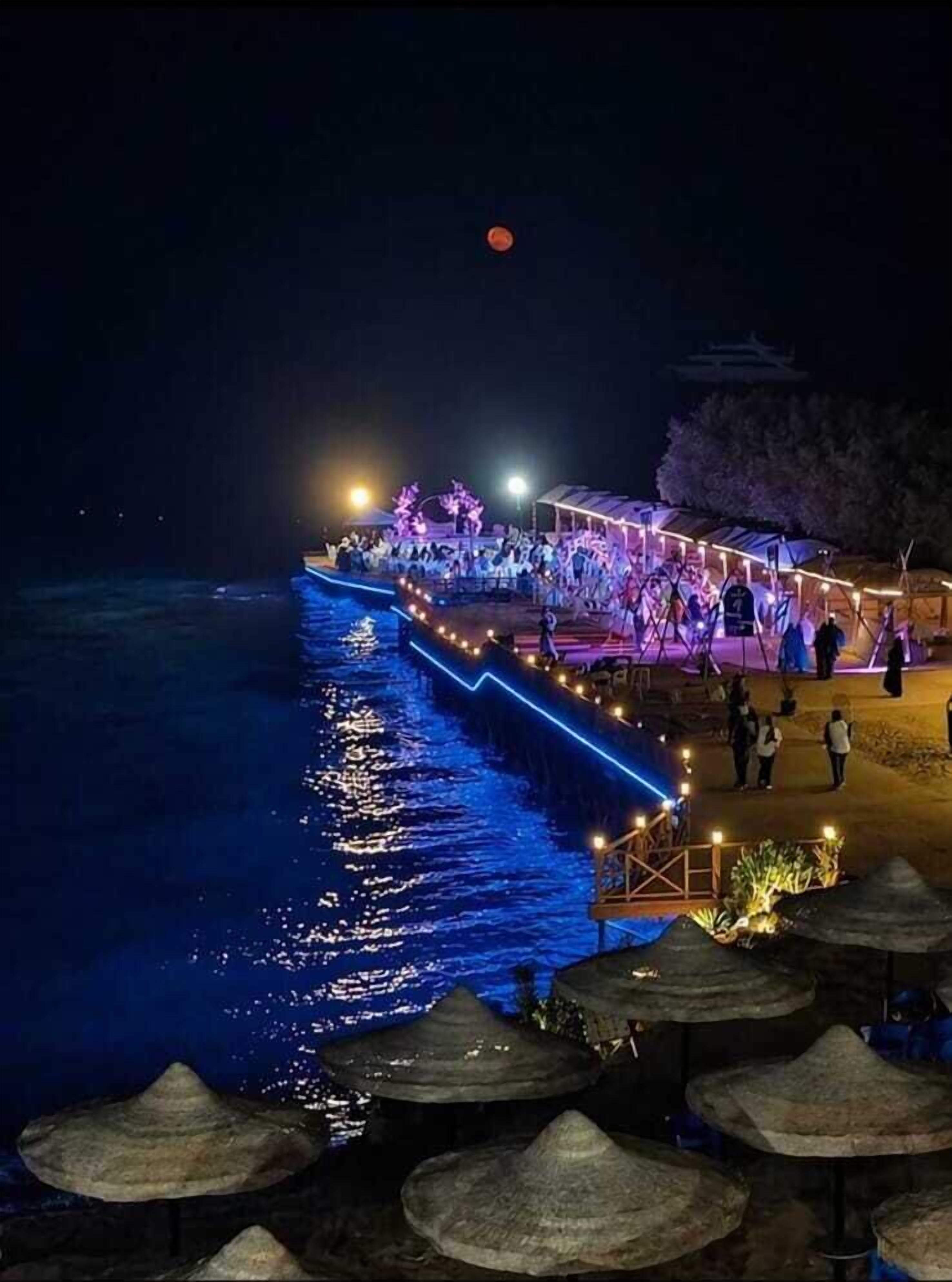 Zona costera con un paseo marítimo, personas y una luna en el cielo.