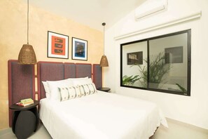 A hotel room with a bed, two framed pictures on the wall, a window with a view, and a small table with a book.