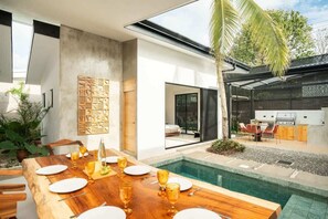 Un espace repas extérieur moderne, doté d’une piscine, d’un barbecue et d’une table en bois dressée pour un repas.