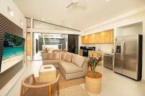 A modern living room with a sectional sofa, a dining area, a kitchen with stainless steel appliances, and a large window with a beach view.