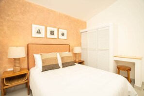 Une chambre à coucher avec un lit, deux tables de chevet avec des lampes, un tabouret en bois, une étagère fixée au mur et une fenêtre avec des stores.