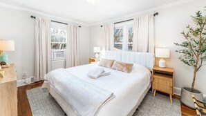 Une chambre d’hôtel bien aménagée, avec un grand lit, deux tables de chevet avec des lampes, une plante en pot et une vue sur les arbres par les fenêtres.