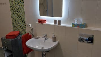 A bathroom with a white sink, a mirror, a towel rack with red towels, a shelf with toiletries, and a shower area.