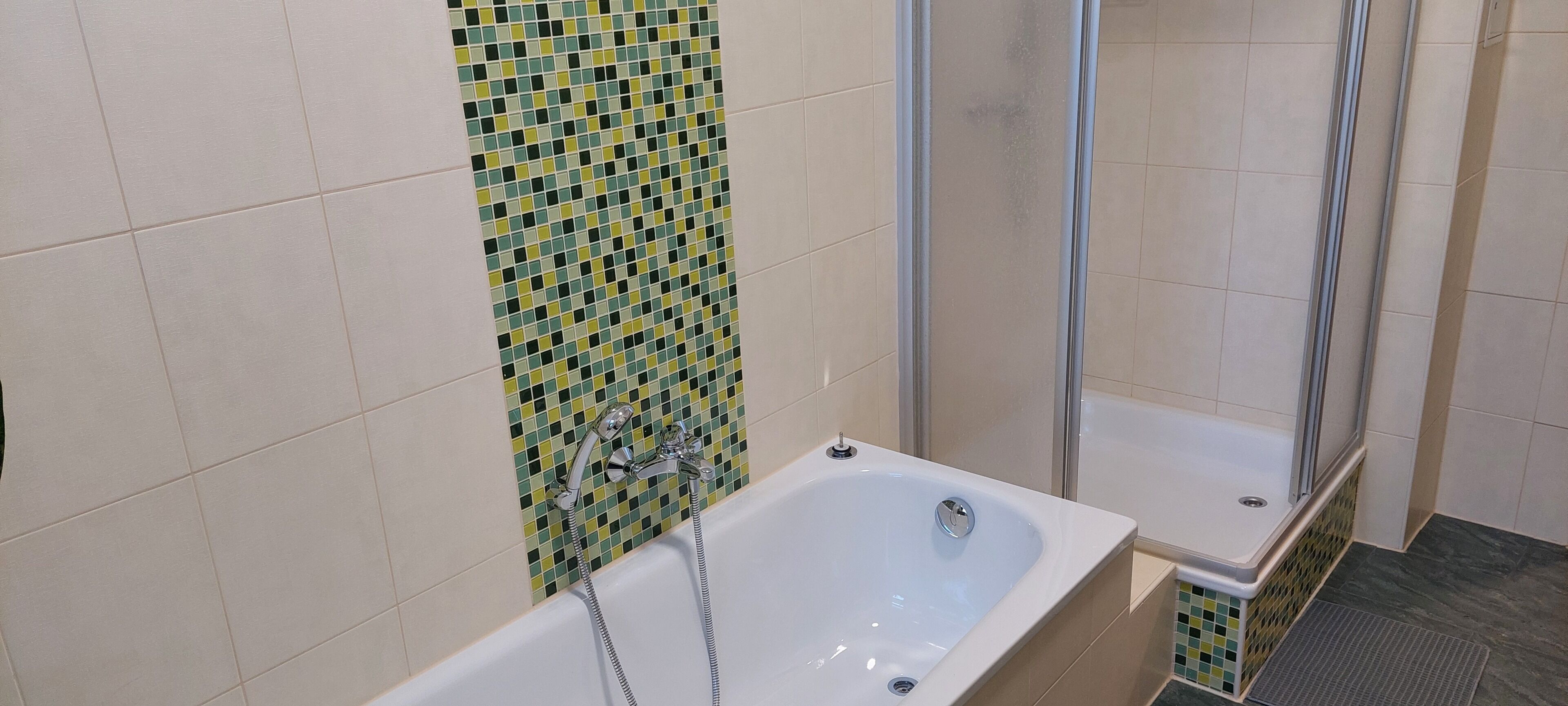 Een badkamer met een bad, een douchegedeelte en een betegelde muur met een groen en geel patroon.