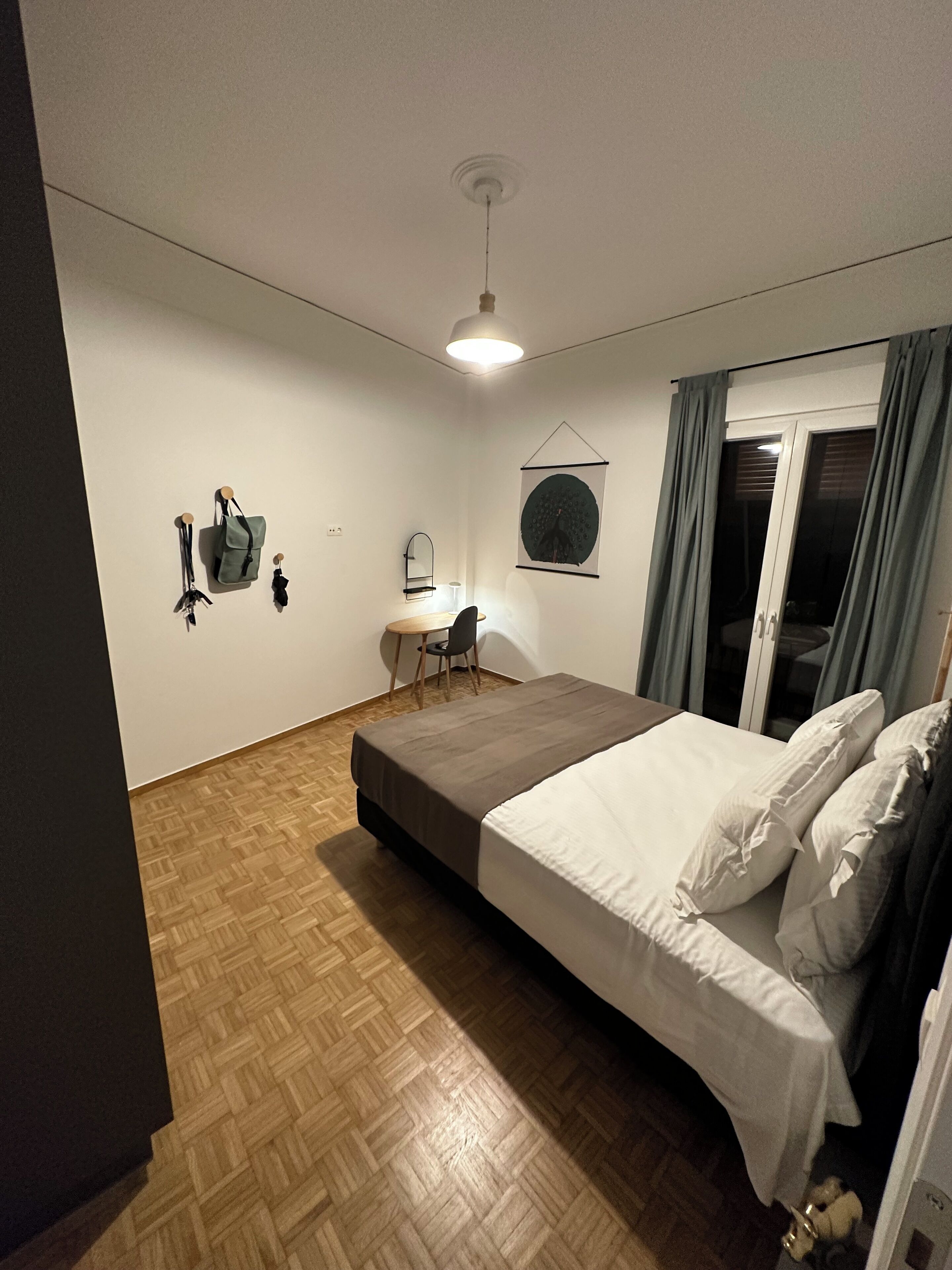 A bedroom with a bed, a desk, a chair, a hanging bag, and a wall-mounted mirror.