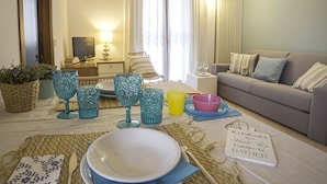 A cozy living room with a dining area set up for a meal. There is a sofa, a television, and a table with plates, glasses, and a bowl. - Bellasirmione - Acquachiara (Sirmione)