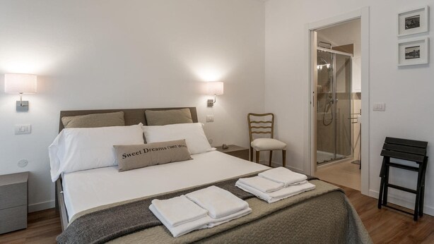 A neatly made bed with white linens and a brown pillow, a bedside table, a chair, and a small table with towels. - Peschierahome - Arilica (Peschiera del Garda)