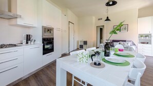 A modern kitchen with white cabinetry, a gas stove, and an oven. There is a dining area with a white table set for two, green placemats, and a vase with flowers. - I Voltoni - PeschieraHome (Peschiera del Garda)