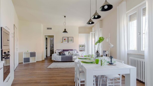 A modern kitchen with white cabinetry, a dining area with a white table and chairs, and a living area with a sofa and decorative pillows. - I Voltoni - PeschieraHome (Peschiera del Garda)