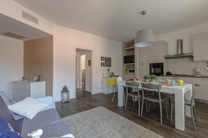 A modern living room with a sofa, a dining table with chairs, a kitchen area with cabinets and appliances, and a hallway leading to another room. - La Rocca - Peschierahome (Peschiera del Garda)