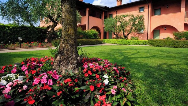 A building with a garden in front, featuring a tree and flower beds. - Bellasirmione - Le Vele (Sirmione)