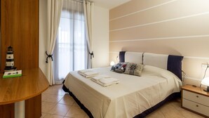 A neatly made bed with pillows and folded towels, a bedside table with a lamp, and a view of the outside through a window with curtains. - Bellasirmione - Le Vele (Sirmione)