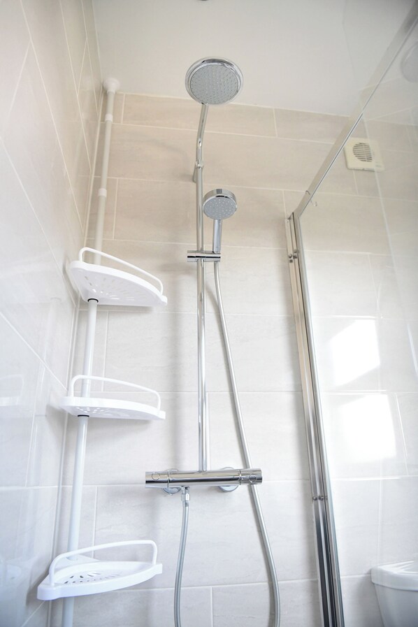 A shower with a handheld showerhead, a wall-mounted faucet, and a handheld showerhead on a shelf.