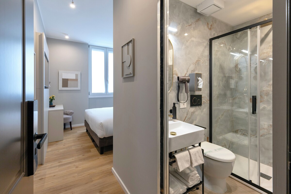A modern hotel room with a wooden floor, a bed, a small desk with a chair, a bathroom with a glass shower enclosure, and a toilet.