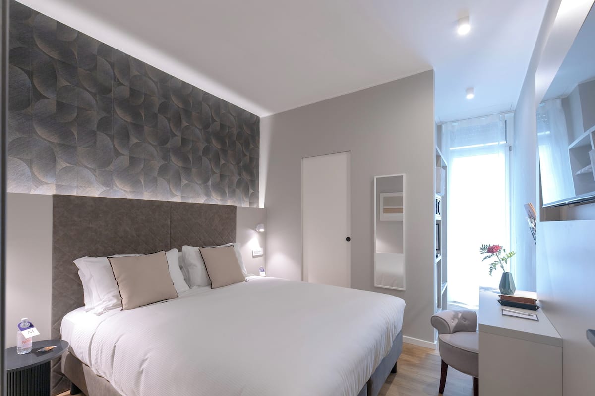Habitación de hotel moderna con una cama grande, cabecera con diseño texturizado, mesita de noche con una botella de agua, una silla y un escritorio con un florero y libros.