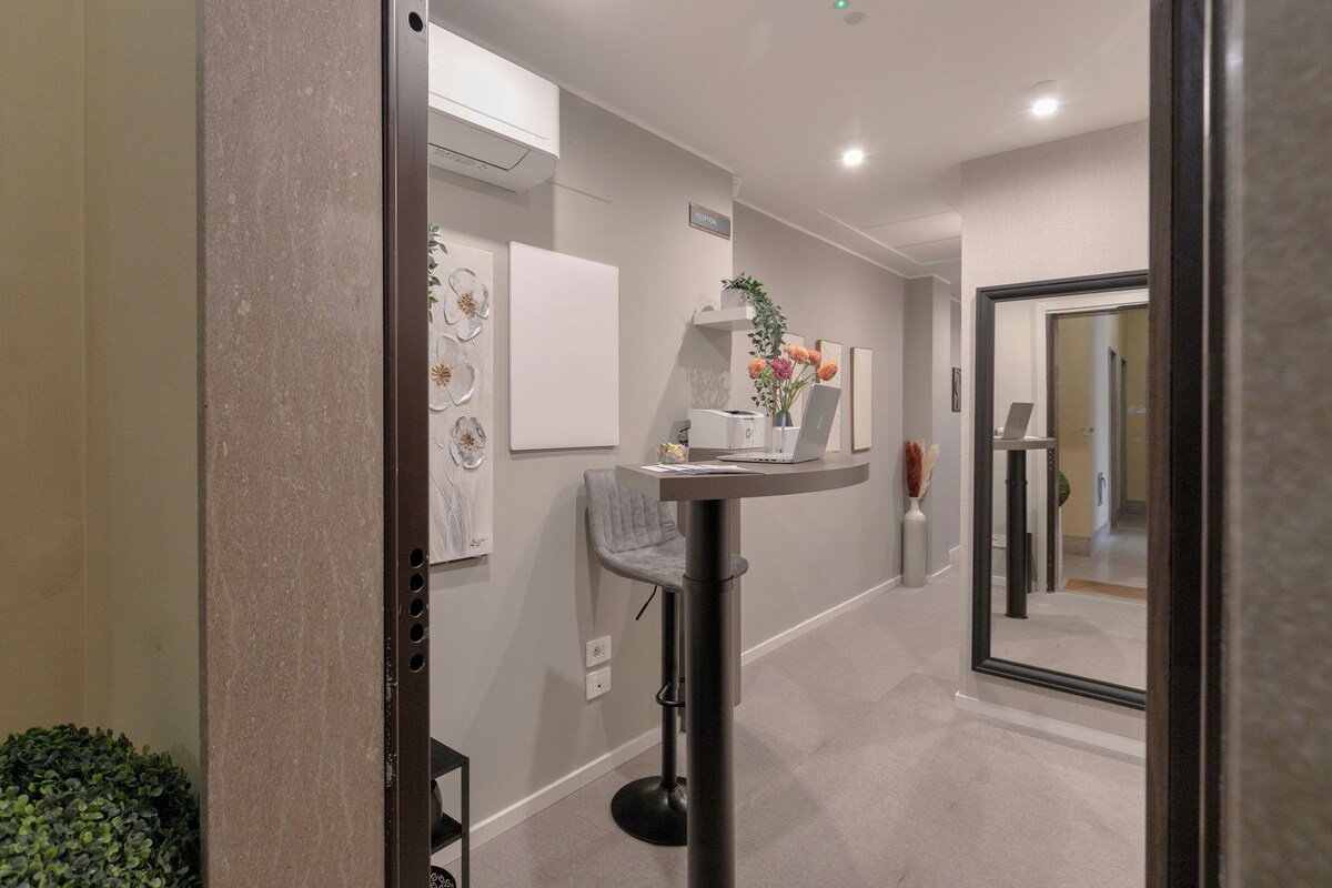 A modern hallway with a round table, a chair, and a laptop. There is a potted plant and a vase with flowers on the table.