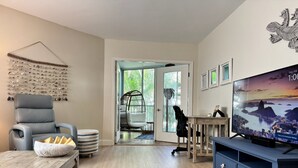 A modern living room with a grey leather sofa, a wooden coffee table, a blue TV stand, and a large window offering a view of the outdoors.