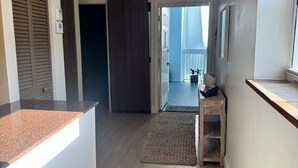 A hallway with a wooden floor, a ceiling light, a door with a dark curtain, and a small wooden stool with a plant.