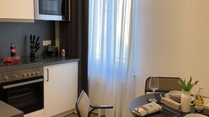 Et kompakt køkken med moderne hårde hvidevarer, et spiseområde med bord og stole, samt et vindue med lette gardiner.