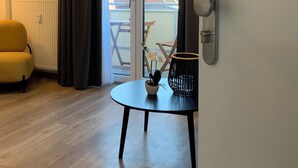 Et værelse med trægulv, en gul lænestol, et bord med en potteplante, og udsigt til en balkon med stol og bord.