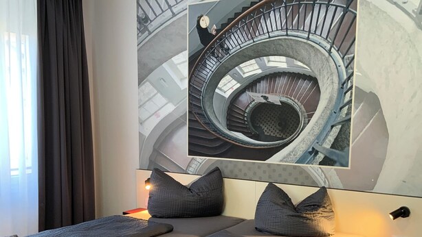 Et moderne hotelværelse med en stor seng, en lille sofa, et maleri af en spiraltrappe og et rødt natbord.