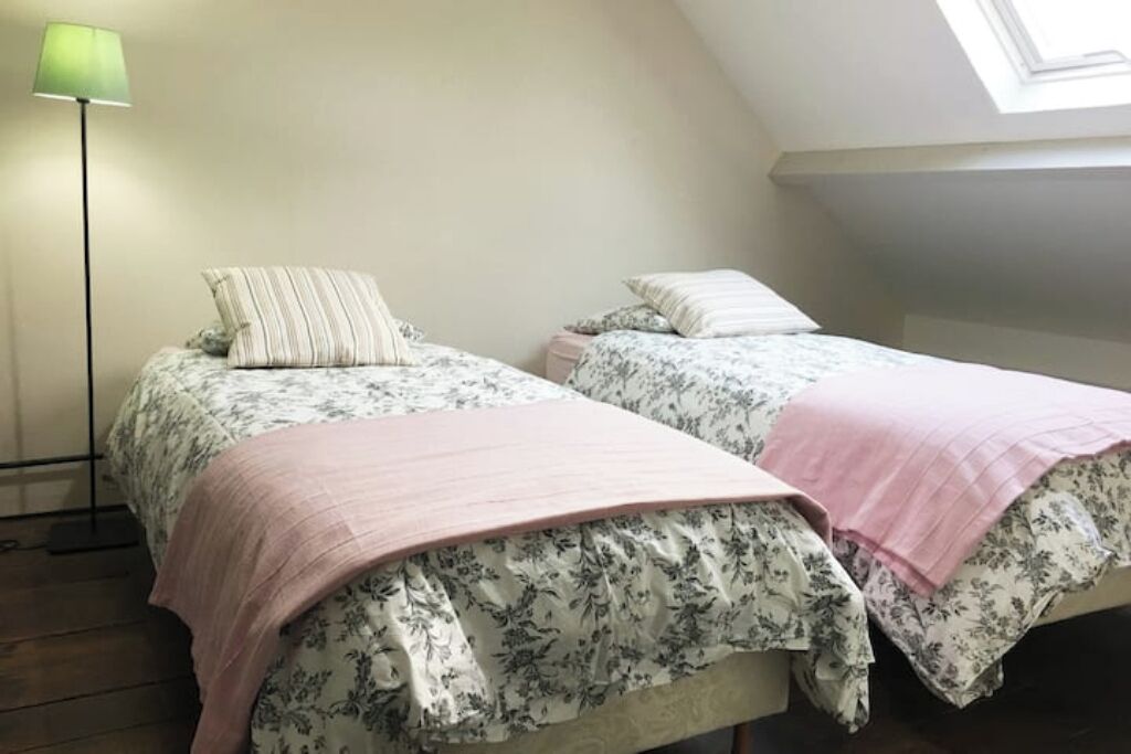 A bedroom with two beds, each with floral bedding and pink throw pillows. A floor lamp is visible on the left side.