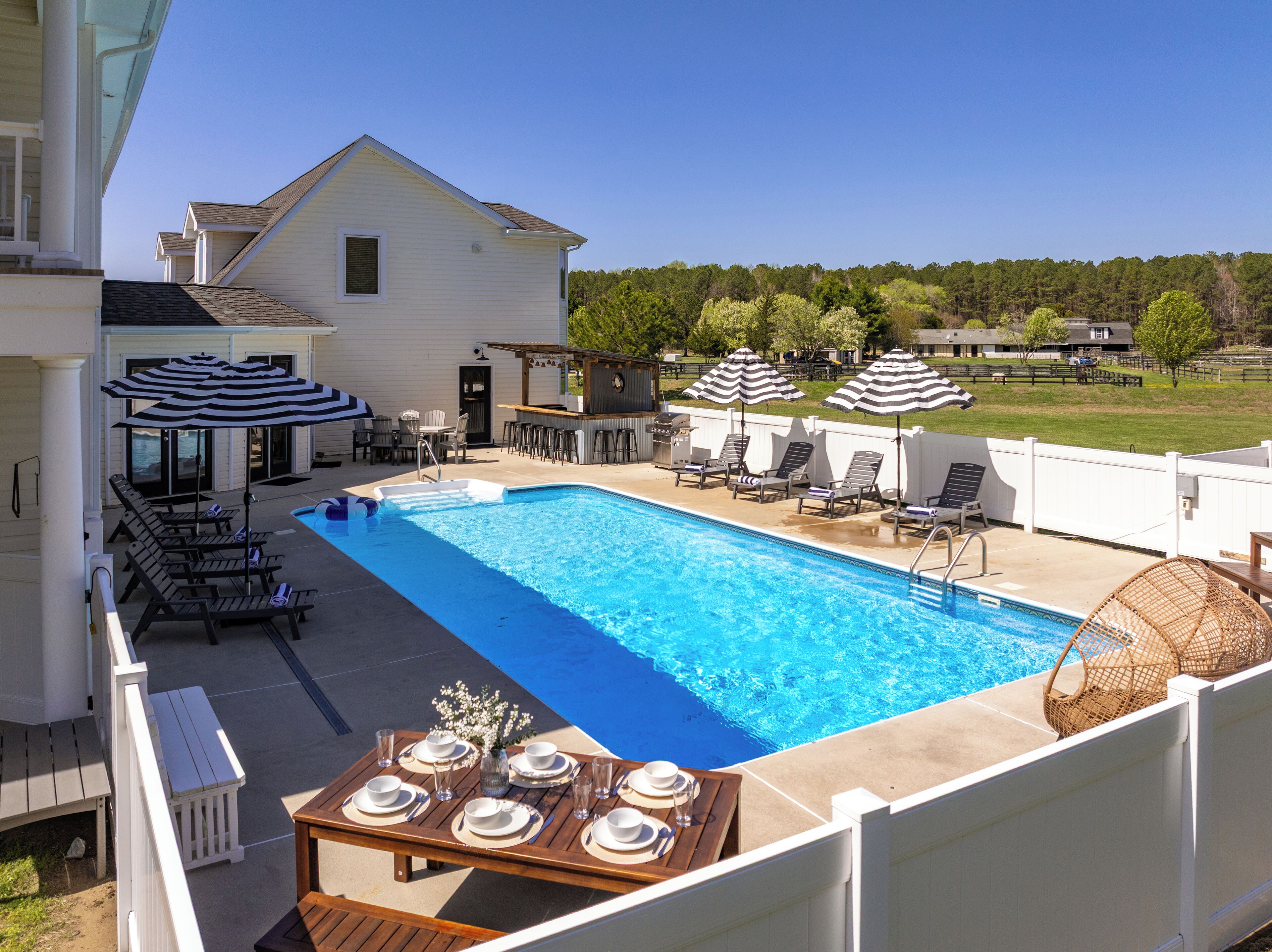 A backyard with a swimming pool, lounge chairs, umbrellas, and a dining table set for an outdoor meal.