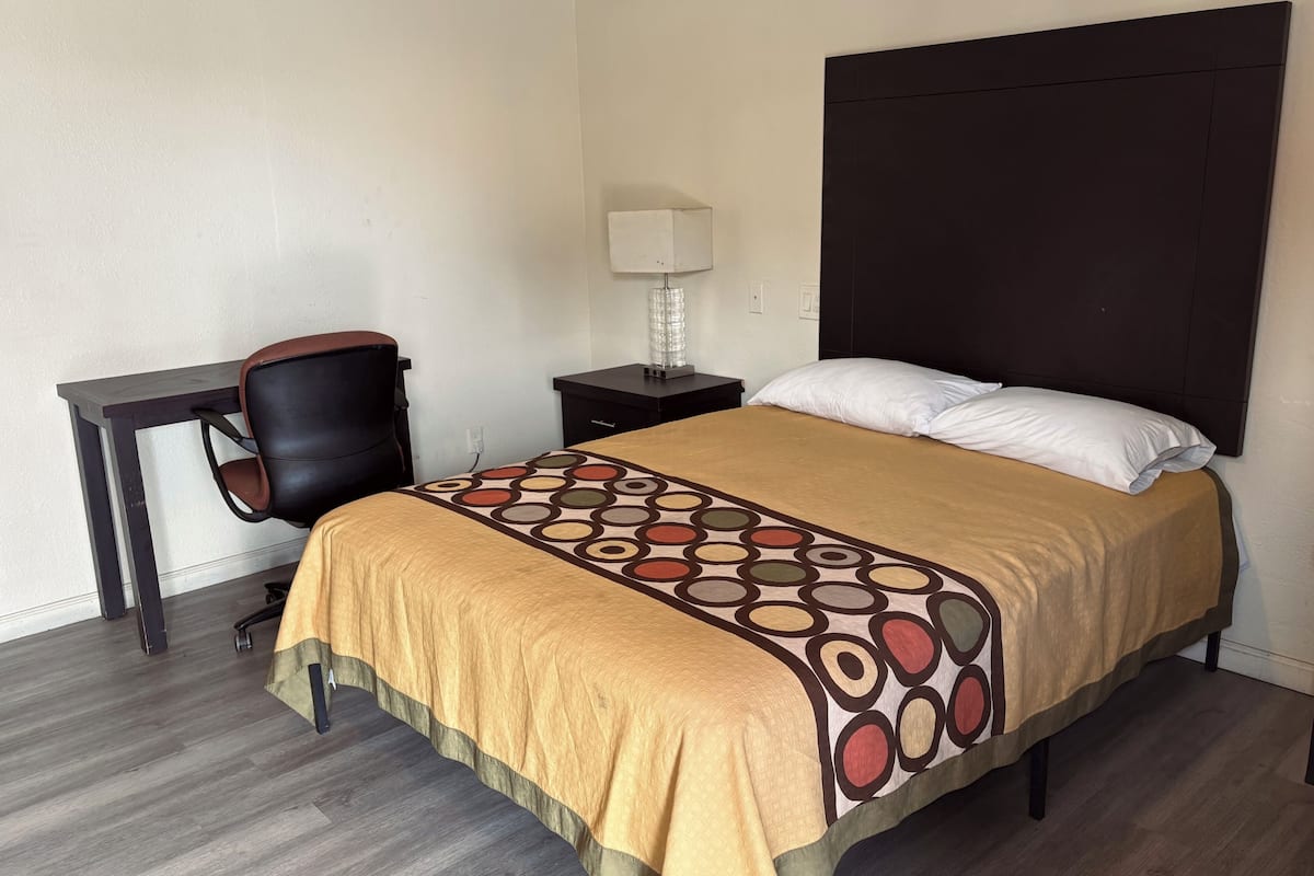A hotel room with a bed, a desk, a chair, a nightstand, and a lamp.