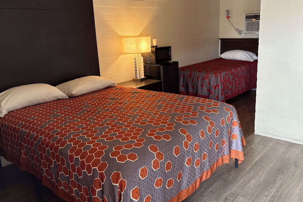 A hotel room with two beds, a nightstand, a lamp, a microwave, and a wall-mounted air conditioning unit.