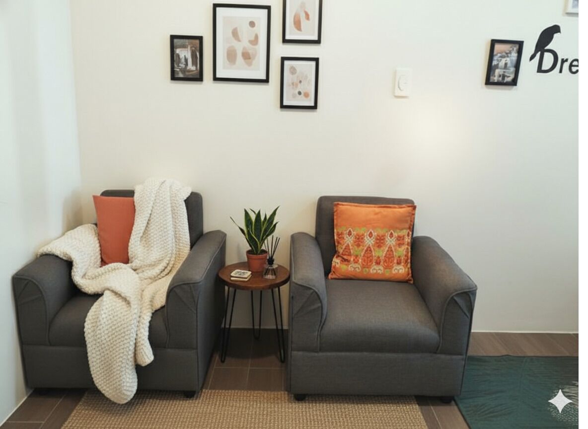 Uma sala de estar com dois poltronas cinzas, uma com um cobertor branco e outra com uma almofada laranja. Há uma mesinha com uma planta e um quadro emoldurado entre as poltronas.