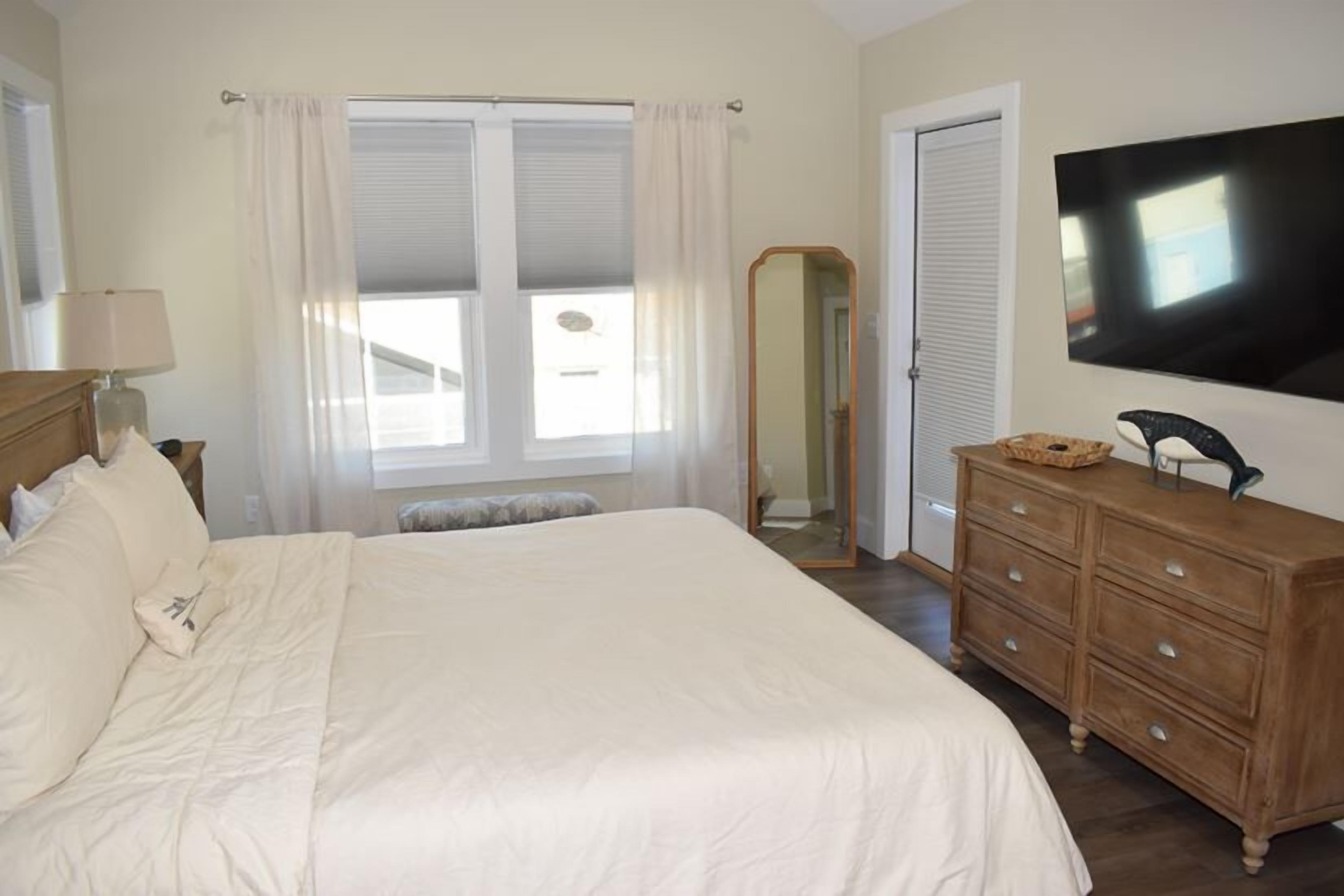 A bedroom with a large bed, a dresser, a mirror, a television, and a window with curtains.