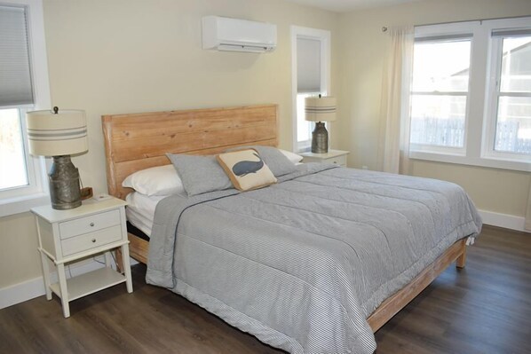A bedroom with a wooden headboard, a grey striped bedspread, two bedside tables with lamps, and a window with curtains.