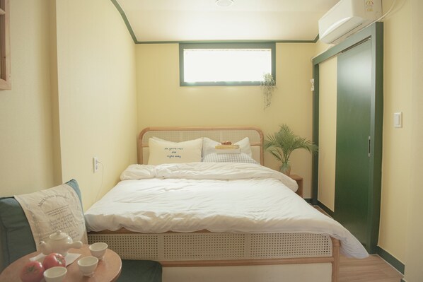 Un dormitorio pequeño y ordenado con una cama individual, una mesita de noche con tetera y tazas, una pequeña silla con cojín y una ventana con planta.