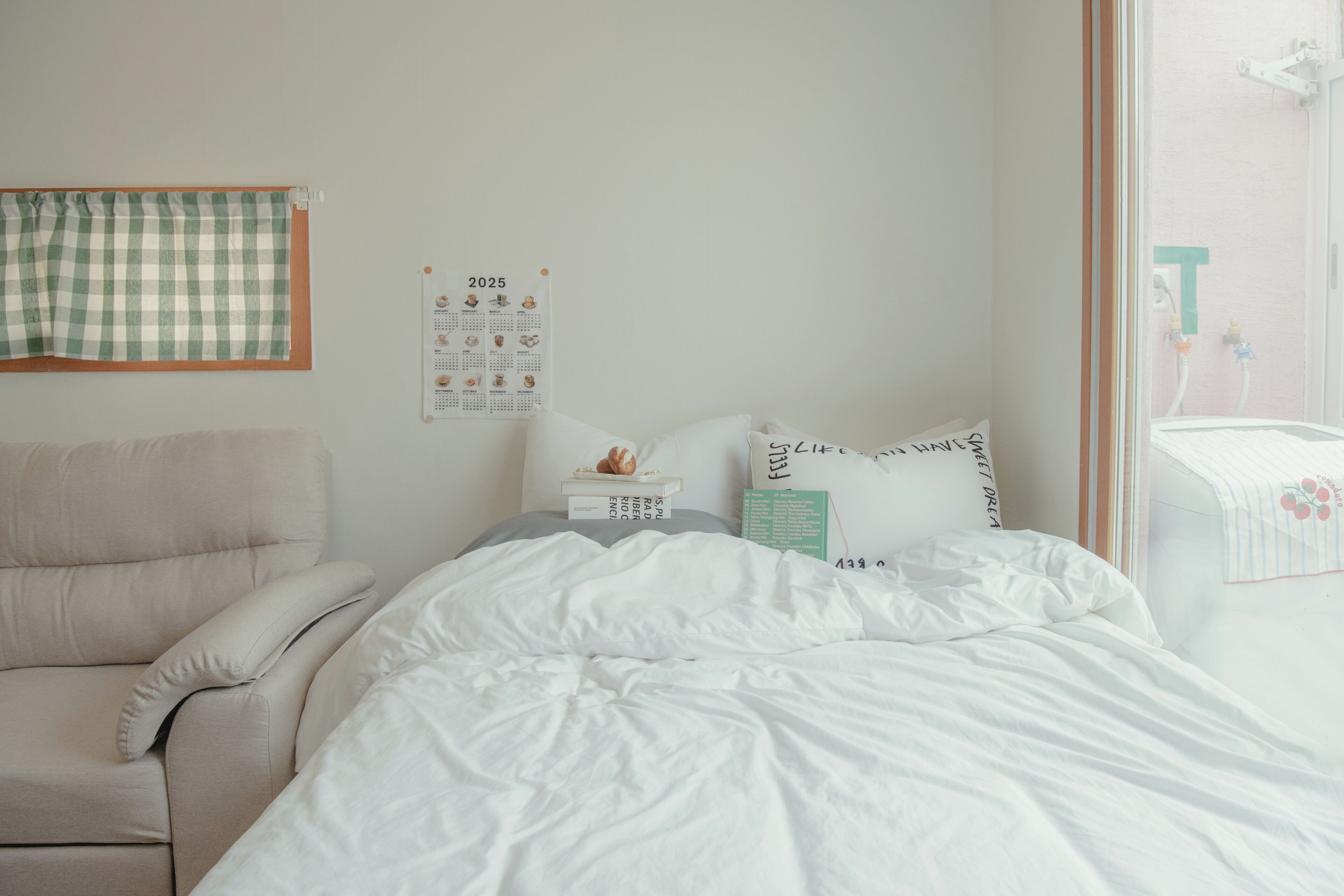 Quarto com cama, cadeira, calendário e janela com vista para um banheiro.