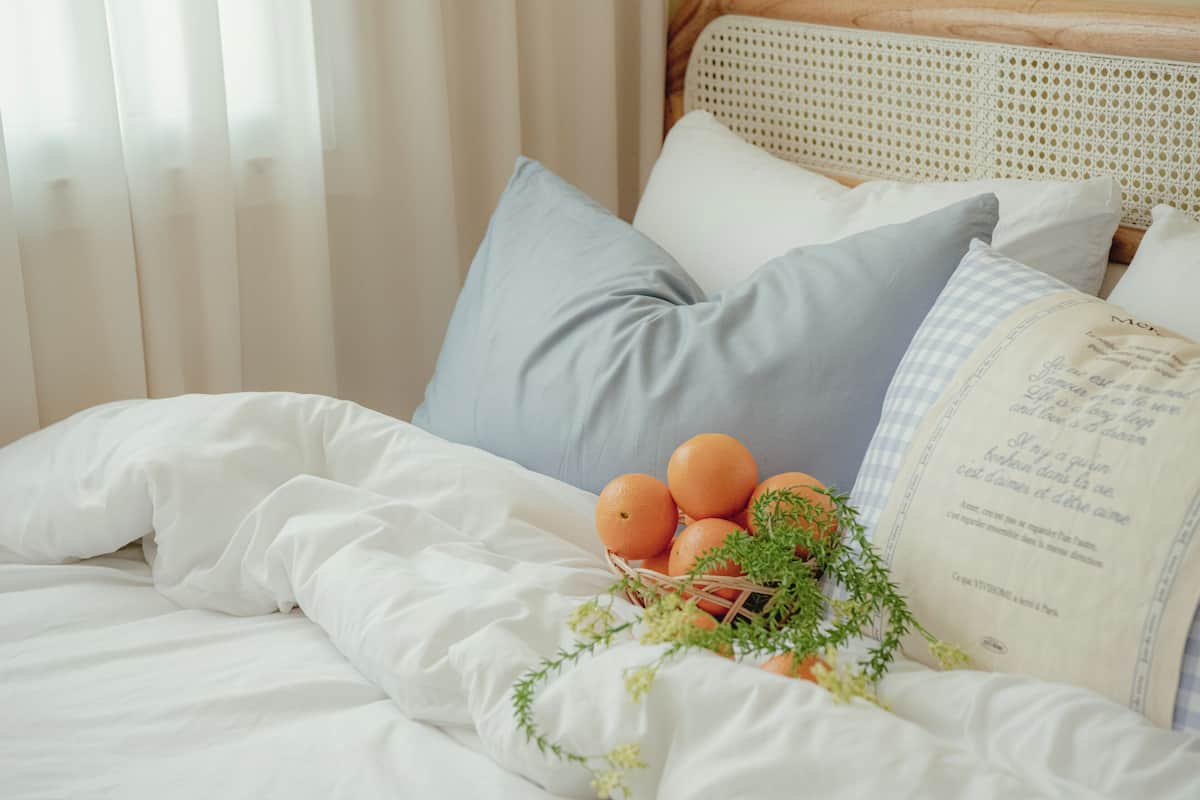A neatly made bed with white bedding, a basket of oranges, and a decorative pillow.