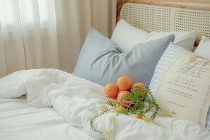 Una cama bien hecha con ropa blanca, una cesta de naranjas y un cojín decorativo.
