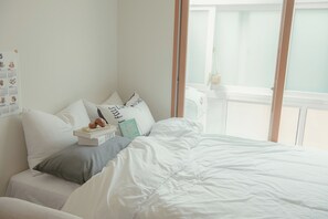 Una cama bien hecha con ropa blanca, una almohada gris y un libro encima. Se ve una ventana con vista a un balcón.