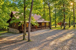 Une cabane en rondins dotée d’un porche et d’une fondation en pierre, entourée d’arbres et de gravier.