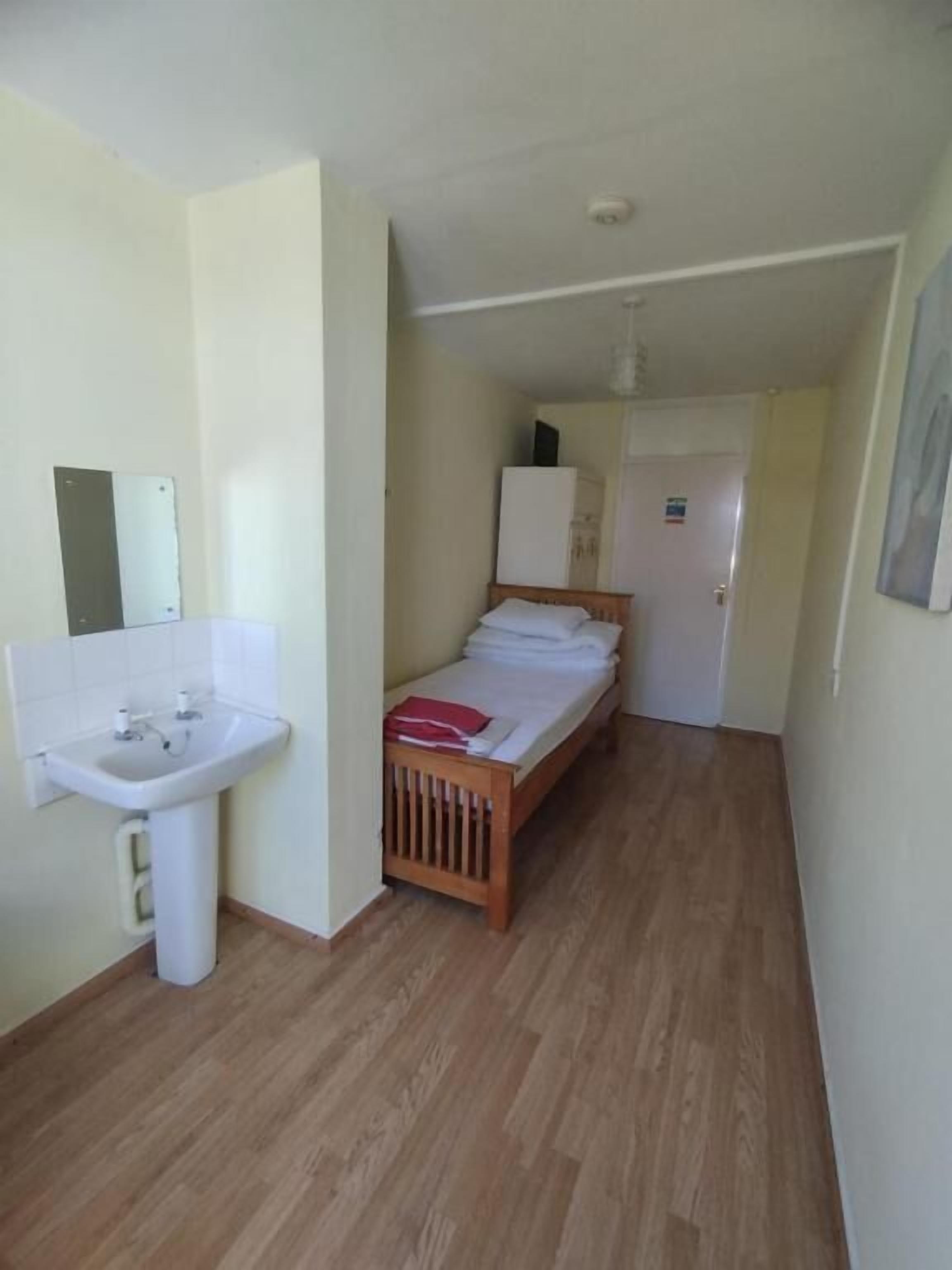 Quarto pequeno e compacto com soalho de madeira, uma cama simples, uma pia branca e uma porta que dá para outro espaço.