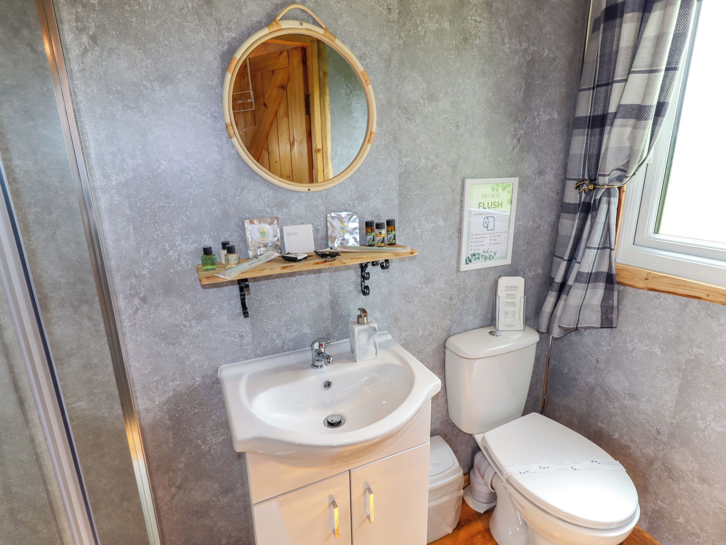A bathroom with a wooden vanity, a sink, a toilet, and a mirror. There is a shelf with toiletries and a 'DO NOT FLUSH' sign.