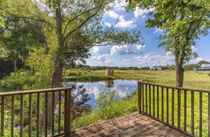 A wooden deck overlooking a calm pond with a fence, surrounded by lush greenery and trees. - Charming 3-bedroom country cottage on a 
Small  pond (Bellville)