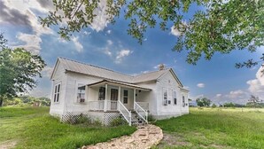 A white wooden house with a porch and steps leading to the front door, surrounded by a grassy field and trees. - Charming 3-bedroom country cottage on a 
Small  pond (Bellville)