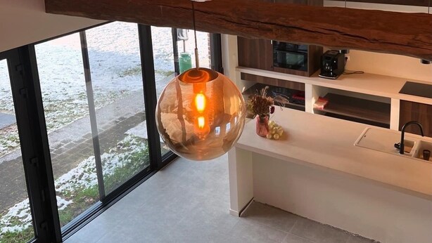 Une cuisine moderne avec un plafond haut et des poutres apparentes, agrémentée de trois lampes suspendues sphériques et d’une grande fenêtre offrant une vue sur l’extérieur.