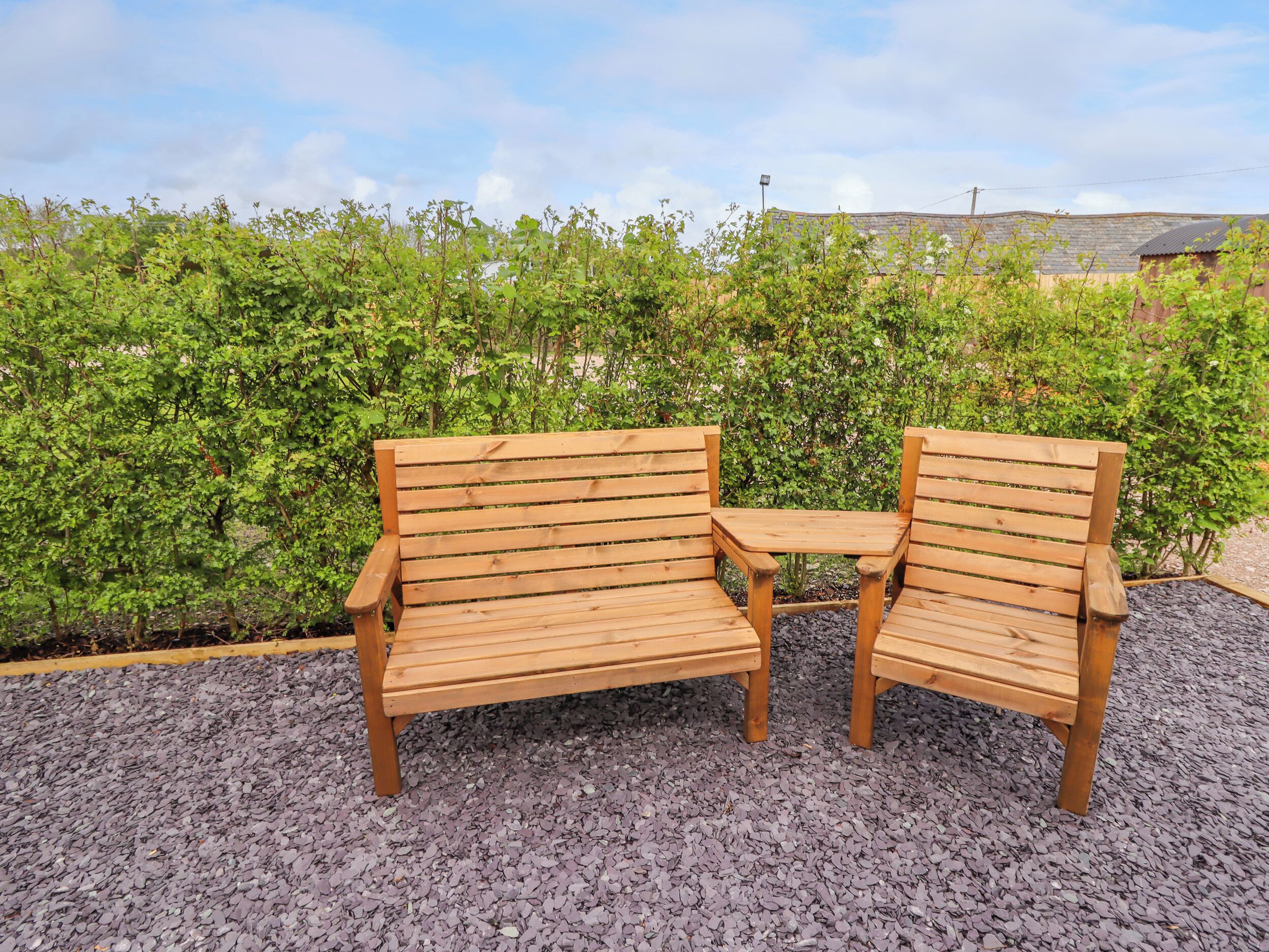 A wooden bench with a small table in front, surrounded by gravel and greenery.
