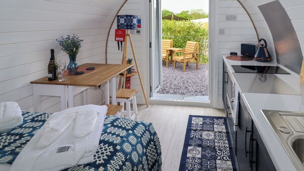 Una habitación acogedora y compacta con una cama, una mesita con una botella y vasos, una silla y vistas a un jardín desde una puerta abierta.
