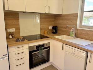 A modern kitchen with white cabinets, a black induction cooktop, a built-in oven, a dishwasher, and a window providing natural light.