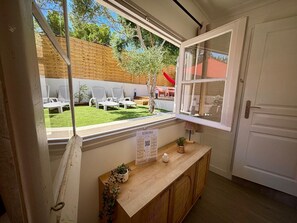 A room with a wooden desk, a window with an open view to a backyard with white chairs and a red slide.